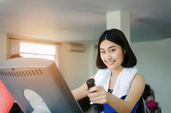 young happy smile asian woman slim body looking into camera and exercising his legs cardio training on bicycle in fitness gym for good healthy at the morning, bodybuilder, lifestyle and sport concept