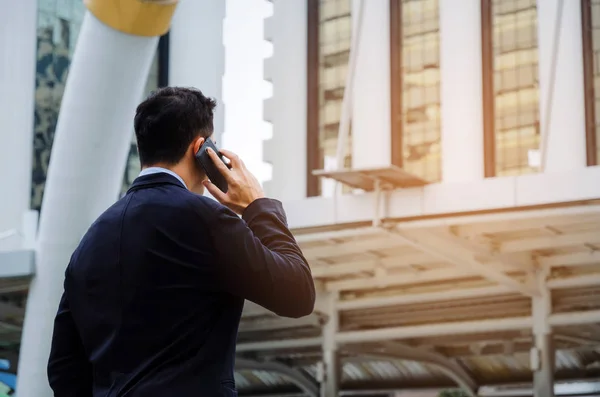 arkadan görünüşü yapı şehir arka plan, Internet ağ bağlantısı, teknoloji iletişim, mali kavram mobil akıllı telefon ile telefon görüşmesi yapma siyah takım elbise giyen genç akıllı iş adamı