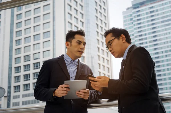 two business asian man talking and reading information about finance news in mobile phone tablet together standing in modern city, network technology, internet, successful, teamwork concept