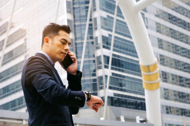 Modern siyah giyen genç akıllı Asya iş adamı uygun saatine bakıyor ve mobil akıllı telefon ile telefon görüşmesi yapma şehir arka plan, ağ bağlantısı, teknoloji iletişim kavramı