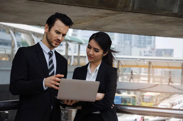 two business asian woman and caucasian man in suit talking and reading information about finance news in laptop computer in modern city, network technology, internet, successful and teamwork concept