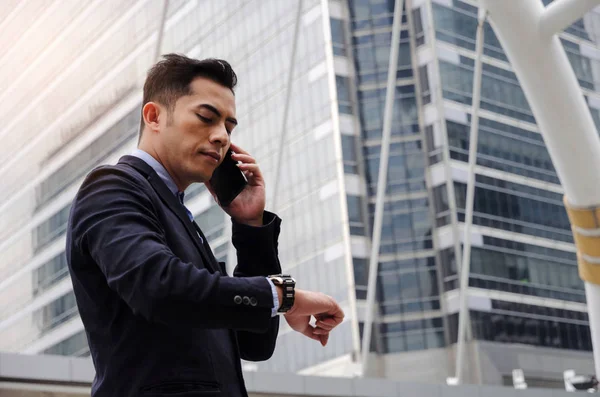 young smart asian business man wearing modern black suit looking at his watch and making phone call with mobile smart phone in city background, network connection, technology communication concept