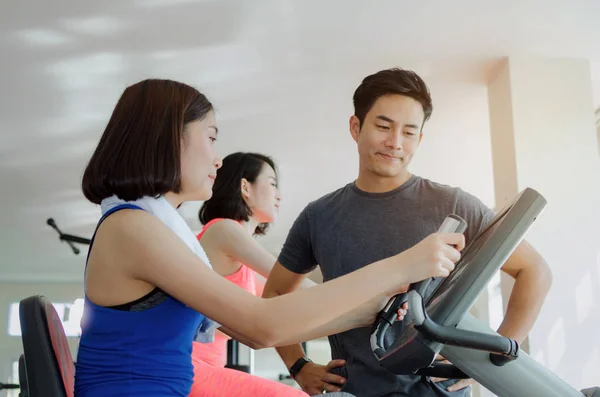 muscular handsome personal trainer man talking with young asian woman slim body with towel and exercising his legs cardio training on bicycle in fitness gym for good healthy, lifestyle, sport concept