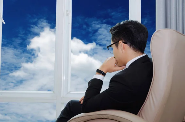 side view of young handsome business man with eyeglasses sitting on sofa chair in modern office with blue sky and cloud in background from window, savings, investment, finance and banking concept