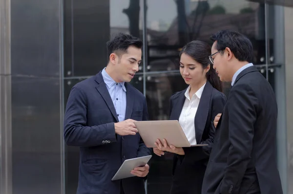 group of business people in suit talking and reading information about finance news in laptop computer together standing in modern city, network technology, internet, successful, teamwork concept