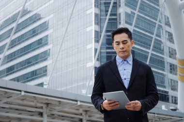 finans haberleri bina şehir arka plan, ağ teknolojisi, Internet, başarılı konsept mobil tablet ile ilgili bilgileri okuduktan modern siyah takım elbise giyen genç yakışıklı Asya iş adamı