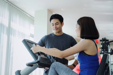 Genç Asyalı kadın ince beden havlu ile ile konuşmak ve onun bacakları kardiyo egzersizi bisiklet için iyi sağlıklı, yaşam, fitness salonunda egzersiz kas yakışıklı kişisel antrenör adam spor kavramı