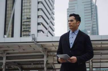 finans haberleri bina şehir arka plan, ağ teknolojisi, Internet, başarılı konsept mobil tablet ile ilgili bilgileri okuduktan modern siyah takım elbise giyen genç yakışıklı Asya iş adamı
