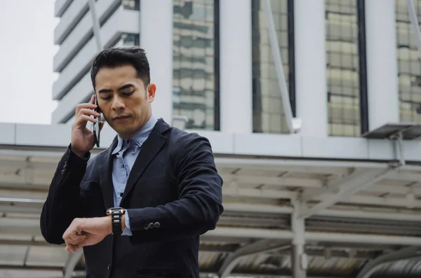 young smart asian business man wearing modern black suit looking at his watch and making phone call with mobile smart phone in city background, network connection, technology communication concept