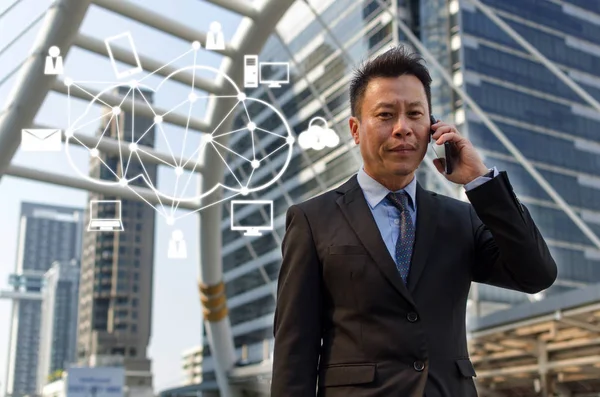 medium aged asian business man wearing modern black suit making phone call with mobile smart phone in city with graphic network icon, investment, technology, internet marketing and successful concept