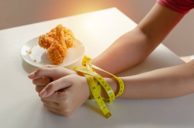 Diyet olsun. Genç kadın sıska elleri sarı mezurayla bağlanmış ve leziz kızarmış tavuk tabağı ev ofisinin mutfağında, abur cubur, sağlıksız yemek, diyet, kilo kaybı konsepti