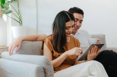 Genç Asyalı çift kahve içiyor ve mobil tablet kullanıyor, ev ofisindeki kanepede film izliyor, internet teknolojisi, sosyal medya, online rezervasyon, eğlence, çevrimiçi alışveriş kavramı