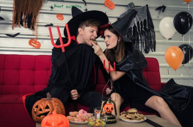 Cadılar Bayramı. Vampir ve cadı kıyafetleri içinde genç beyaz bir erkek ve kadın birlikte tatlı yiyecekler yiyorlar ve Cadılar Bayramı partisinde kanepede oturuyorlar evde, aşk, tatil ve cadılar bayramı festivali konsepti