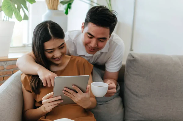 아시아인 커플은 커피를 마시고 휴대용 태블릿을 사용하고 집에서 소파에서 온라인 — 스톡 사진