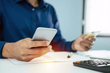 Mavi gömlekli adam güvenlik kodunu dijital cep telefonuyla giriyor ve evdeki masasında sahte kredi kartı ve hesap makinesiyle ödeme yapıyor, online alışveriş, elektronik ödeme, dijital teknoloji konsepti