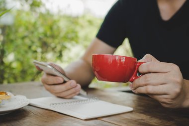 İş adamı elinde kırmızı kahve fincanıyla kafede dijital cep telefonuyla, kendini karantinada, evden çalışma, dijital pazarlama, iş finansmanı, ağ teknolojisi kavramı ile rahat bir şekilde çalışıyor.