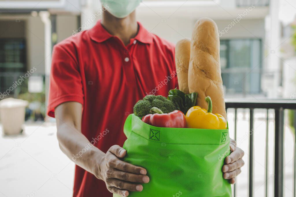 food delivery service man with protection face mask in red uniform holding fresh food set bag waiting for customer at door home, healthy food, express delivery, food delivery, online shopping concept
