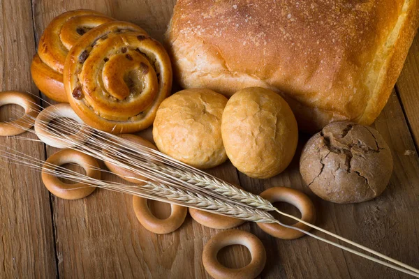 Different types of fresh bread and wheat spikes on old wooden ta — Stock Photo, Image