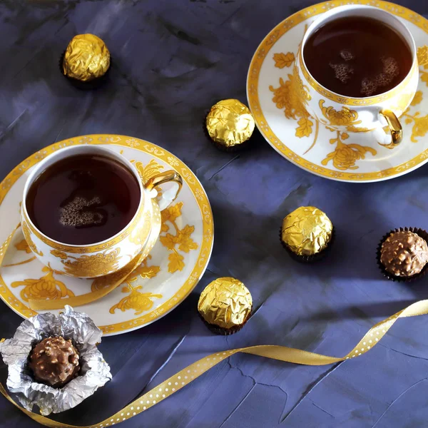 Tea ceremony, tea party. Two tea cups of gold color with black tea, candy, chocolate on a lilac color background.