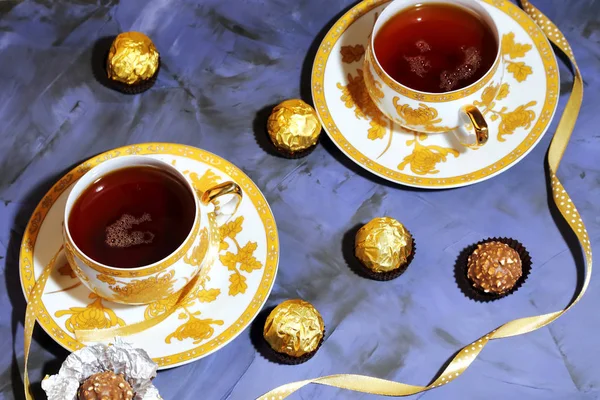 Teezeremonie, Teeparty. zwei goldene Teetassen mit schwarzem Tee, Süßigkeiten, Schokolade auf fliederfarbenem Hintergrund. — Stockfoto
