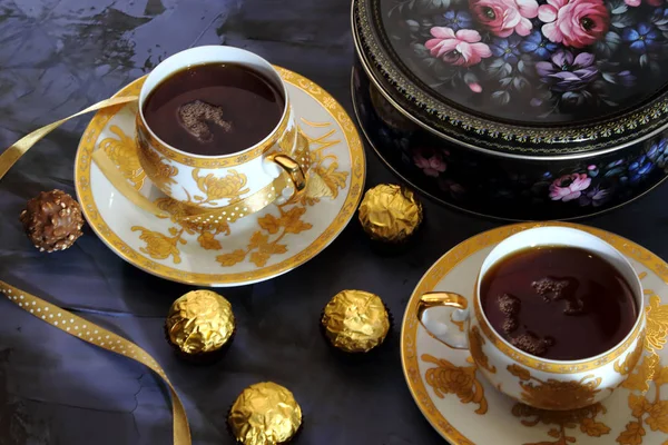 Ceremonii parzenia herbaty, tea party. Dwie filiżanki herbaty złoty kolor czarna herbata, cukierki, czekolady i pudełko z plików cookie na tle w kolorze Lila. — Zdjęcie stockowe