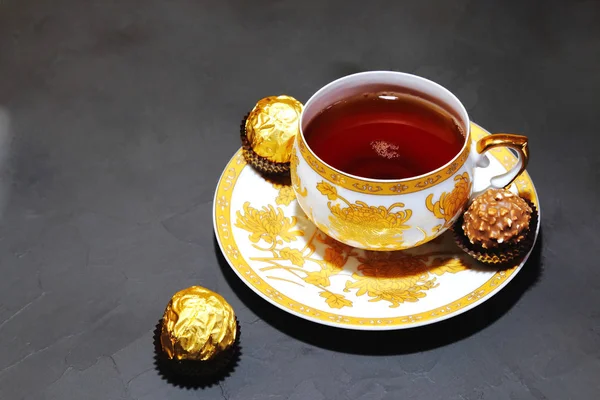 Abendliche Teeparty auf grauem Hintergrund. Porzellan goldene Tasse mit Tee und Dessert Schokoladenbonbons. — Stockfoto