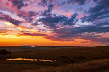 Parlak renkli dramatik manzara. Akşam günbatımında gökyüzü, mavi, mor, turuncu fırtınadan önce gök gürültülü bulutlar, tarlalar ve çayırlar, güneş ufukta batıyor.