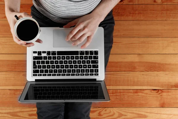 Un portátil y una taza de café en las manos de un hombre sentado en un suelo de madera. Vista desde arriba, copiar espacio . — Foto de Stock