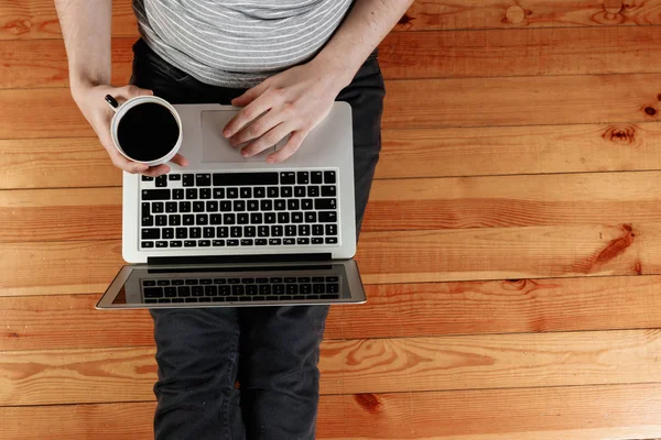A laptop and a cup of coffee in the hands of a man sitting on a wooden floor. View from above, copy space. — Stock Photo, Image