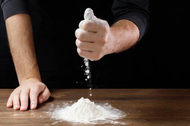 Aşçı yakın çekim el hamur siyah bir arka plan üzerinde pişirmek için masanın üzerine un dökün.