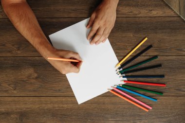 Sanatçı yakın çekim, elinde bir kalem kahverengi ahşap bir masa üzerinde dağınık renkli kalemler yakınındaki boş kağıdın üzerinde tutar. Yukarıdan görüntülemek.