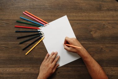 Sanatçı yakın çekim, elinde bir kalem kahverengi ahşap bir masa üzerinde dağınık renkli kalemler yakınındaki boş kağıdın üzerinde tutar. Yukarıdan görüntülemek.