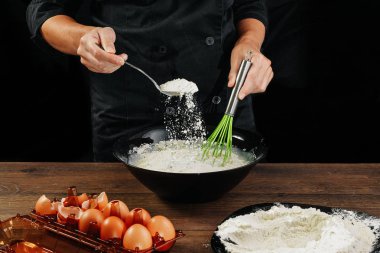 Erkek elinde Şef siyah mutfak eşyaları ahşap kahverengi tabloda un bir tabak içine dökülen. Şef hamur kneads. Siyah arka plan. Yemek yemek kavramı. Yakın çekim. Kopya alanı.