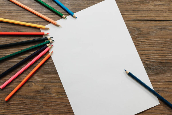 Руки художника крупным планом, держит карандаш над чистым листом бумаги возле разбросанных цветных карандашей на коричневом деревянном столе. Вид сверху
.