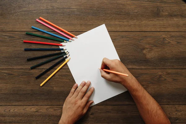 Sanatçı yakın çekim, elinde bir kalem kahverengi ahşap bir masa üzerinde dağınık renkli kalemler yakınındaki boş kağıdın üzerinde tutar. Yukarıdan görüntülemek.