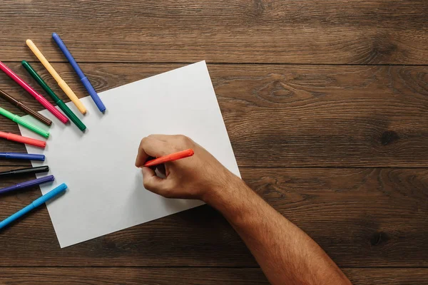 Sanatçı yakın çekim, elinde bir flamaster dağınık renkli keçe bahşiş kahverengi ahşap bir masa üzerinde yakınındaki boş kağıdın üzerinde tutar. Yukarıdan görüntüleyin. Kopya alanı.