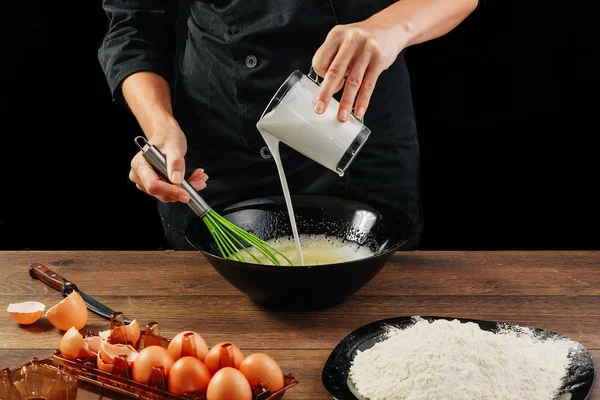 Erkek elinde Şef bir plaka üzerinde bir ahşap kahverengi masa siyah bir kase içine süt dökülen. Şef hamur kneads. Siyah arka plan. Yemek yemek kavramı. Yakın çekim. Kopya alanı.