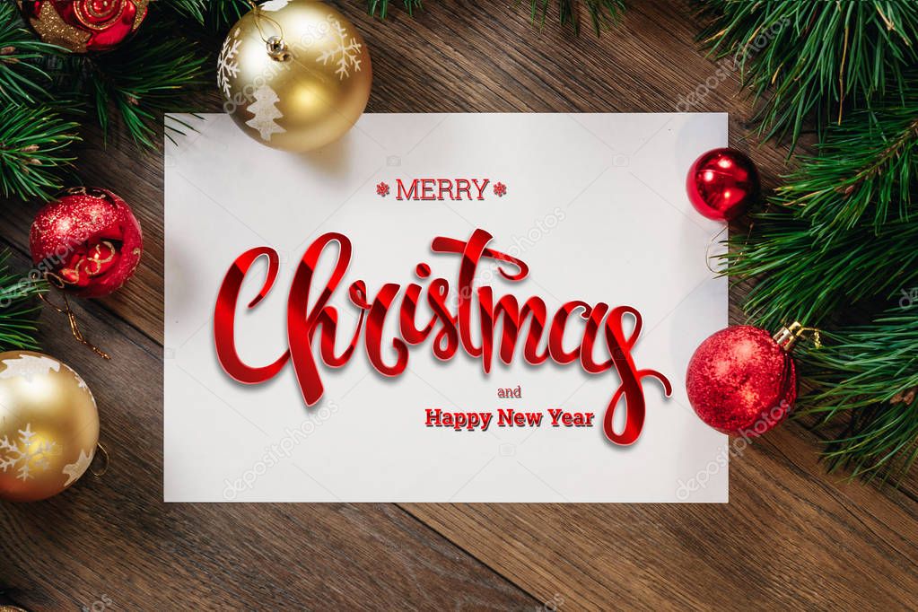 The inscription of Merry Christmas and Happy New Year, green spruce branches, decorations, A4 leaf on a wooden brown table. Christmas card, holiday background. Mixed media.