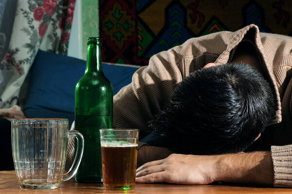 Пьяный мужчина со стеклом и бутылкой алкогольного напитка сидит за столом
