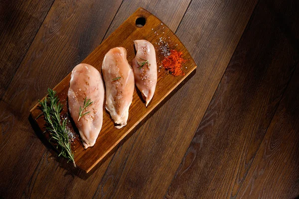 Raw chicken breast, pepper, rosemary on a cutting board, on a wooden background, top view. Raw chicken meat for cooking. Delicious balanced nutrition concept. Copy space