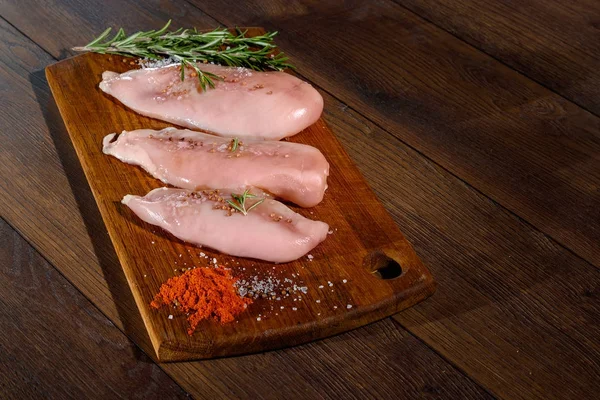 Raw, chicken fillet on a cutting board, a kitchen board, rosemary, pepper, salt, seasoning. The concept of a recipe for cooking chicken.