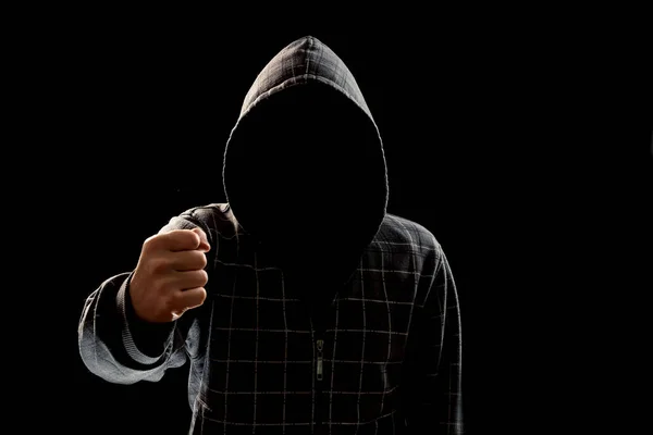 Silhouette of a man in a hood on a black background, his face is not visible, showing a fist in the camera. The concept of a criminal, incognito, mystery, secrecy, anonymity.