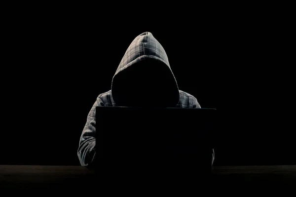 Silhouette of a man in a hood on a black background, his face is not visible, he sits at the computer. The concept of a criminal, incognito, mystery, secrecy, anonymity.