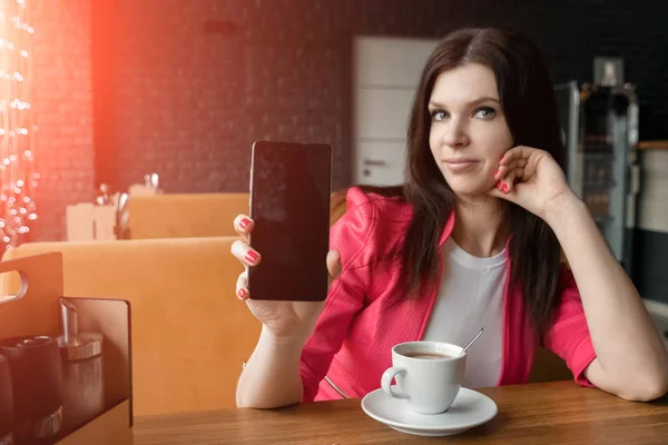 Ung flicka visar telefonen i kameran sitter i café vid bord. Ett träbord, en kopp kaffe. — Stockfoto