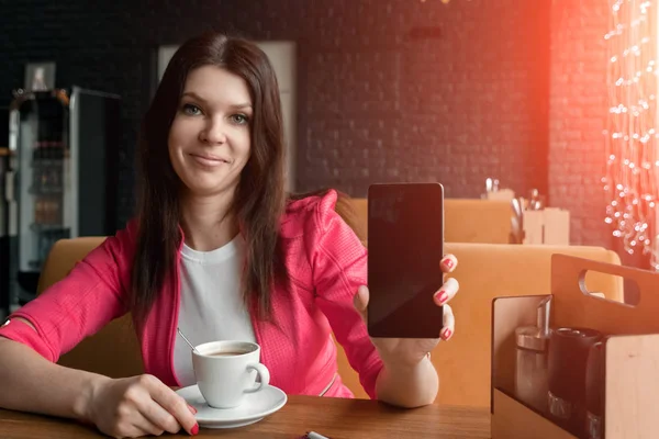 Chica joven muestra el teléfono en la cámara sentado en la cafetería en la mesa. Una mesa de madera, una taza de café . — Foto de Stock