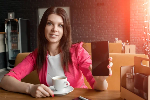 Chica joven muestra el teléfono en la cámara sentado en la cafetería en la mesa. Una mesa de madera, una taza de café . — Foto de Stock