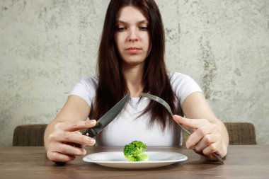 sad young brunette woman dealing with anorexia nervosa or bulimia having small green vegetable on plate. Dieting problems, eating disorder. clipart
