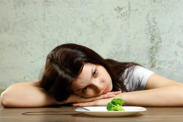 Traurige junge brünette Frau, die mit Magersucht oder Bulimie zu tun hat und kleines grünes Gemüse auf dem Teller hat. Ernährungsprobleme, Essstörungen. — Stockfoto