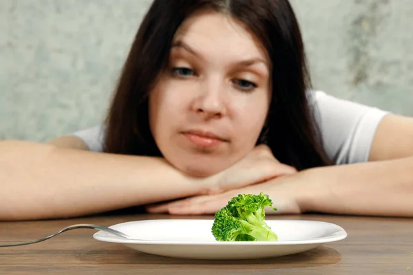 Traurige junge brünette Frau, die mit Magersucht oder Bulimie zu tun hat und kleines grünes Gemüse auf dem Teller hat. Ernährungsprobleme, Essstörungen. — Stockfoto