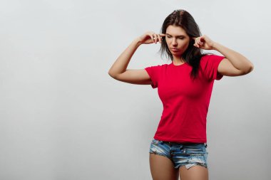 Genç, güzel bir kız onun parmaklarıyla kulaklarını durdurur. Hafif bir arka plan üzerinde izole. Farklı insan duyguları, yüz ifadesi, tutum, algı, beden dili, tepki duyguları.